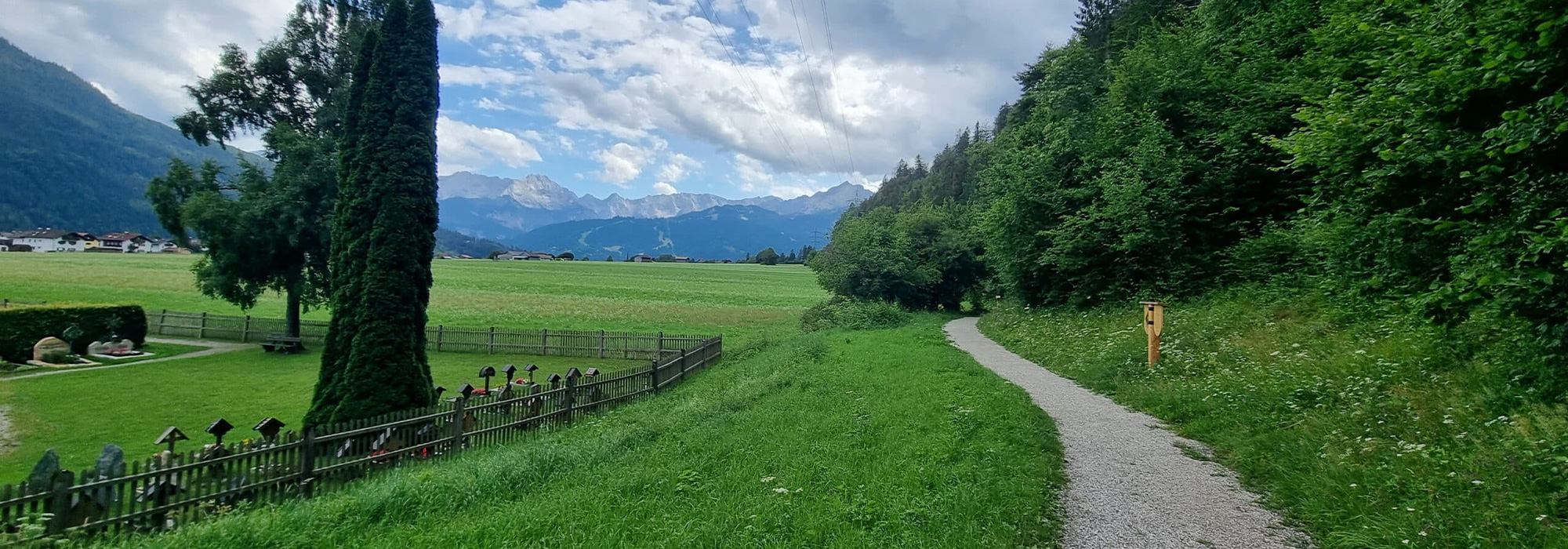 Ein schmaler Schotterweg, rechts davon Wald und hohes Gras, links davon Wiese und der Friedhof von Farchant. In der Ferne das Wettersteinmassiv
